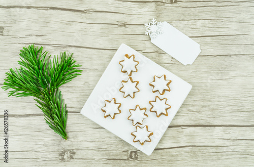Kleiner quadratischer weißer Teller mit Weihnachtsplätzchen, Zimtsternen, einem Tannenzweig und einem weißen Schild, das beschriftet werden kann, Untergrund aus Holz, Textfreiraum