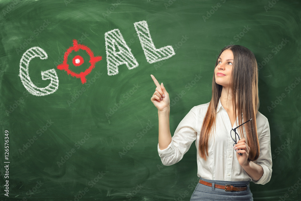 Young woman holding her glasses and pointing finger at word 'goal' written on green chalkboard wall.
