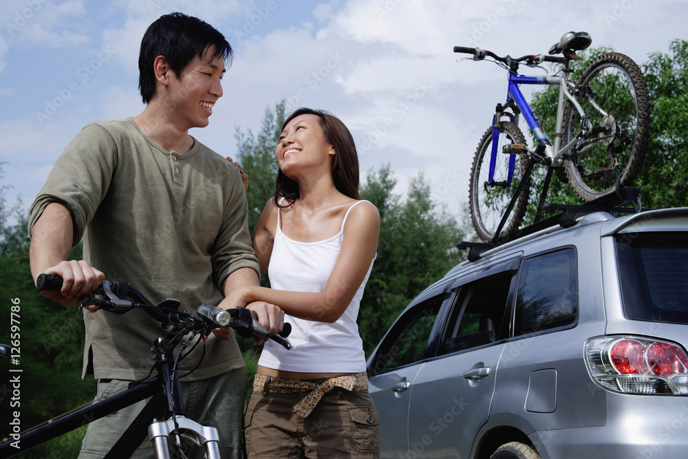 Obraz premium Man on a bike, woman standing next to him, Sports Utility Vehicle in the background