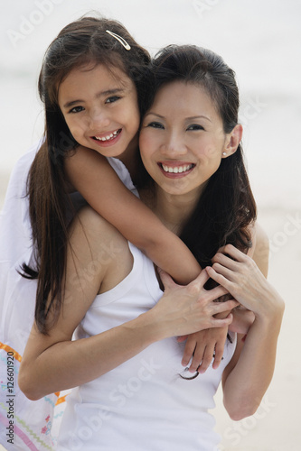 Daughter behind mother with arms over shoulders, smiling, hugging, on the beach