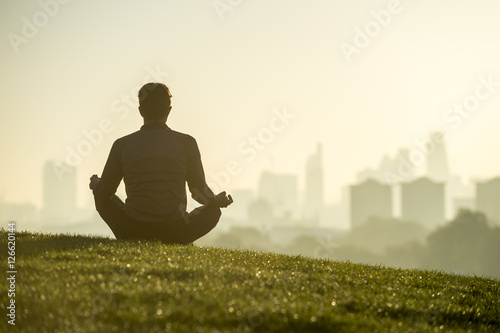 Silhouette of a man sitting in the lotus position meditating on the grassy top of Primrose Hill in front of a misty golden sunrise view of the London city skyline