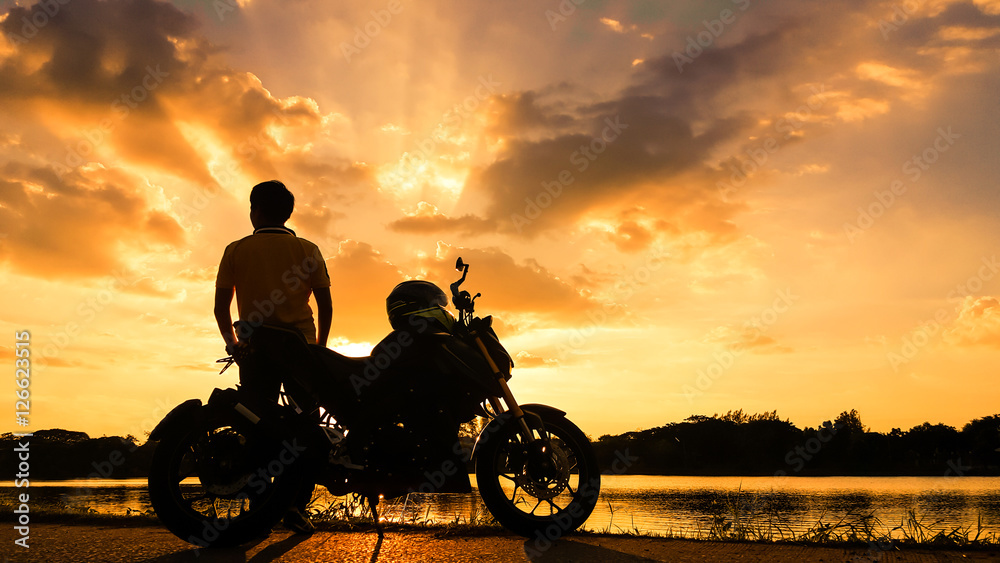 Naklejka premium Sylwetka rowerzysta z jego motocyklem obok naturalnego jeziora i piękne niebo zmierzchu.