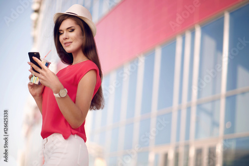 Young beautiful girl in a fashionable hat, red shirt and nice long hair drinking coffee outdoors in the summer in the city and looking at the phone