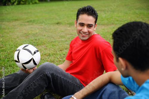 Two men sitting on grass, one holding soccer ball