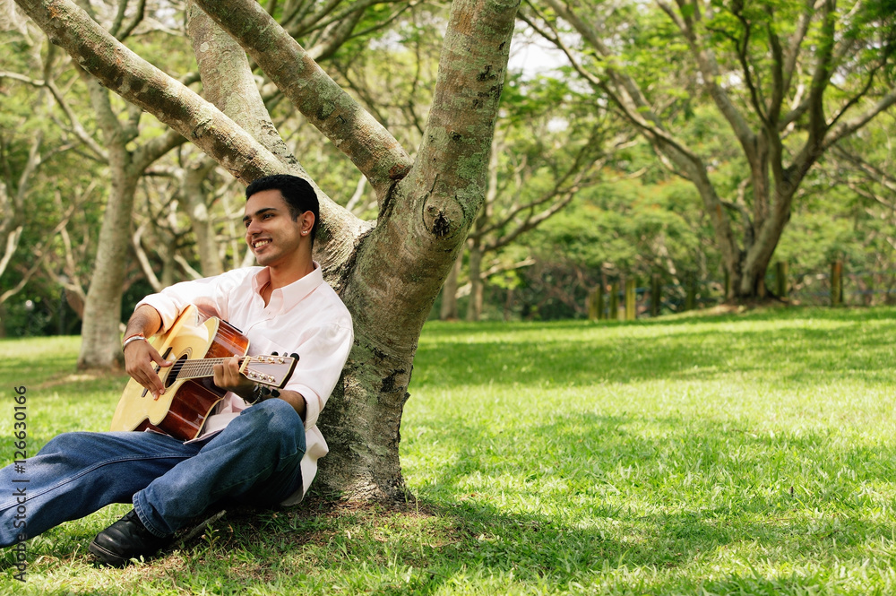 Young man sitting under tree, playing guitar Young man sitting under tree, playing guitar