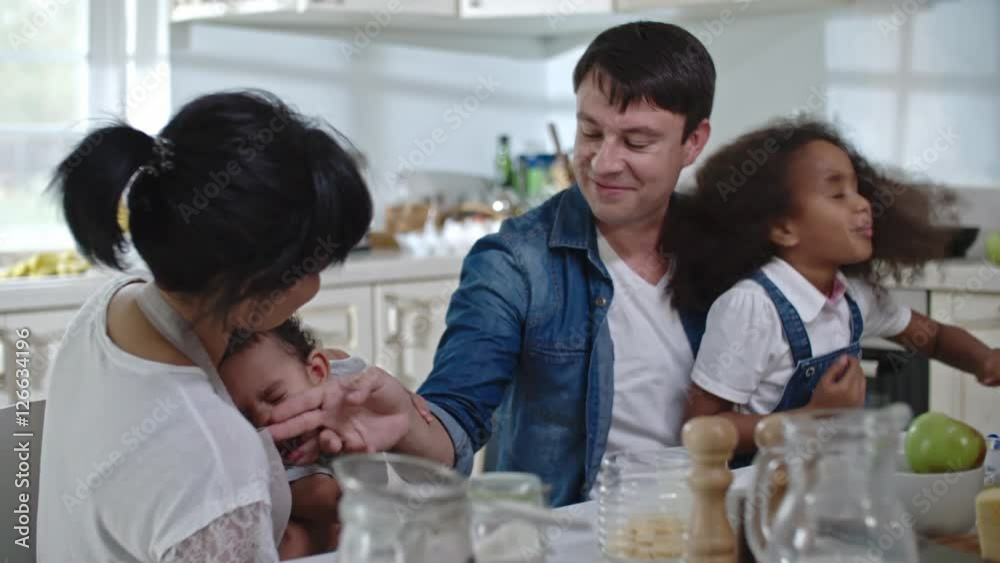 Slow motion of a father wiping milk from face of his little son in the kitchen, cute daughter sitting on lap of her dad, shaking her head with her thick curly hair flying Slow motion of a father wiping milk from face of his little son in the kitchen, cute daughter sitting on lap of her dad, shaking her head with her thick curly hair flying