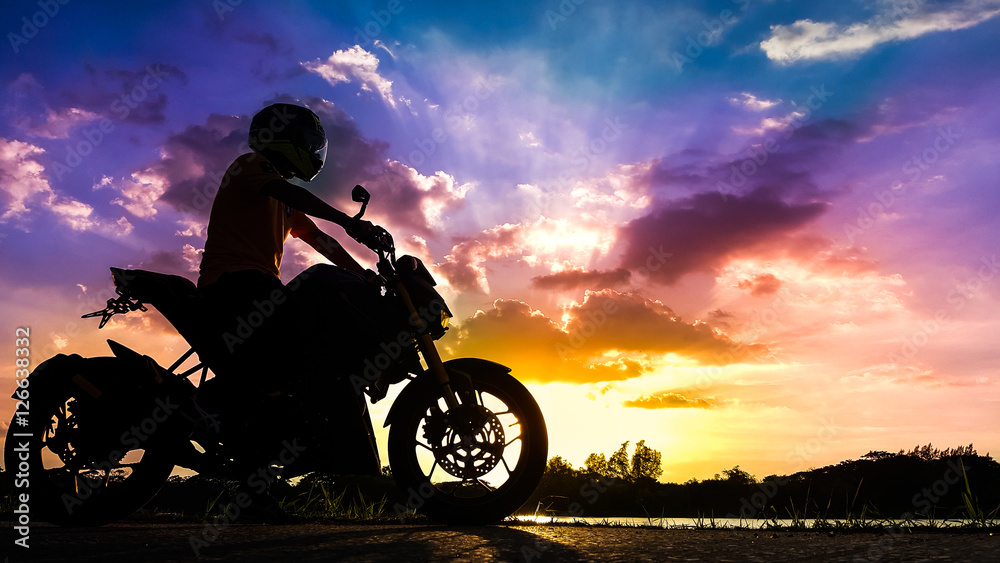 Fototapeta premium Sylwetka rowerzysty ze swoim motocyklem obok naturalnego jeziora i pięknego nieba o zmierzchu.