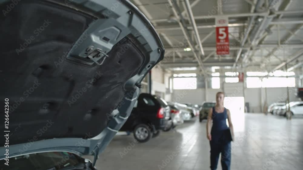 Tilt down of woman in blue uniform walking to car in the repair garage and using laptop for auto diagnostics Tilt down of woman in blue uniform walking to car in the repair garage and using laptop for auto diagnostics