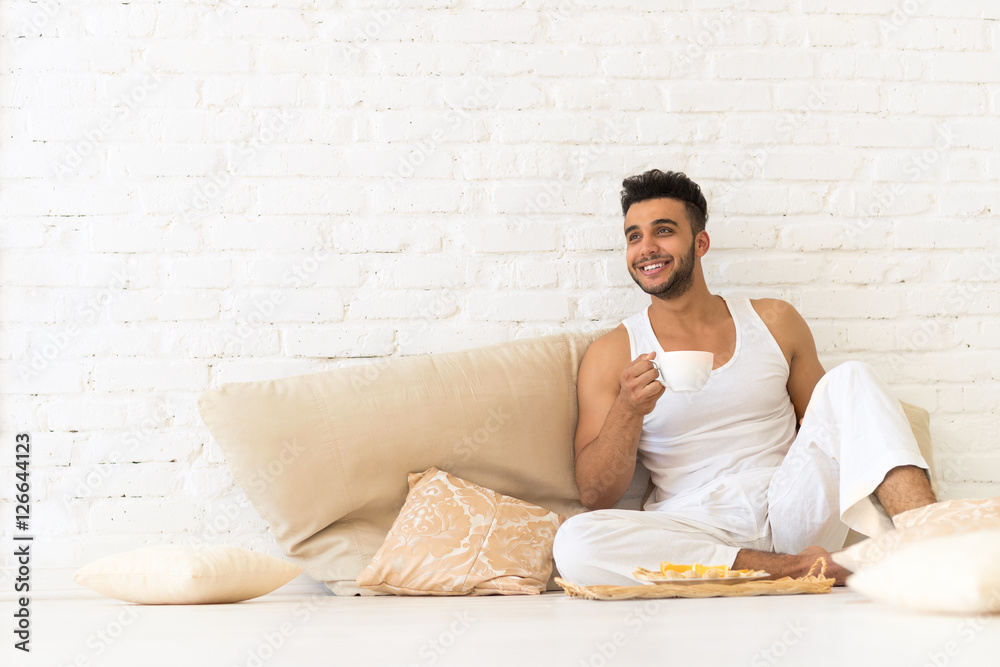 Young Hispanic Man Sit On Pillows Floor, Happy Smiling Guy With Breakfast Tray Look Copy Space Bedroom Over White Brick Wall Young Hispanic Man Sit On Pillows Floor, Happy Smiling Guy With Breakfast Tray Look Copy Space Bedroom Over White Brick Wall