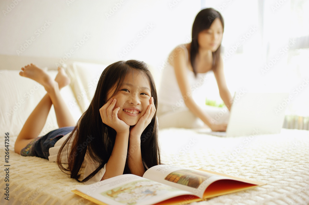 Mother using laptop, daughter lying down with book open in front of her Mother using laptop, daughter lying down with book open in front of her