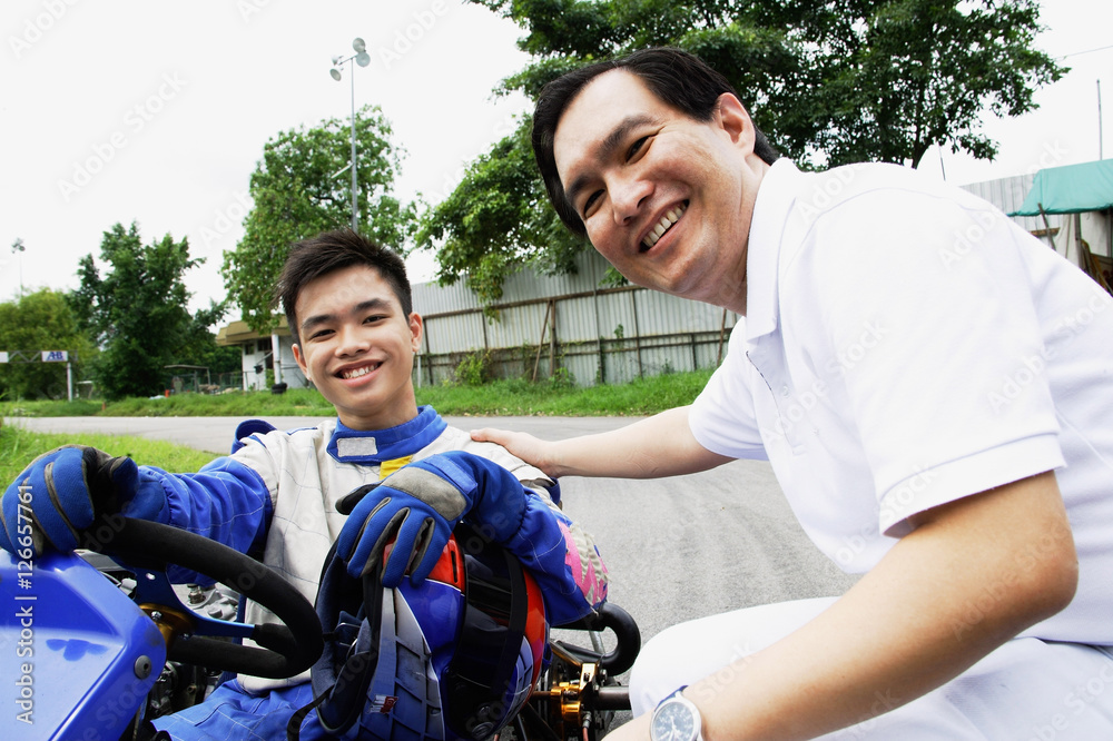 Young man in go-cart, father crouching down next to him, both looking at camera
