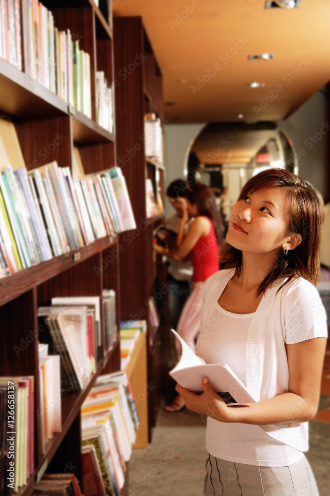 Young woman at bookstore, holding book, looking up