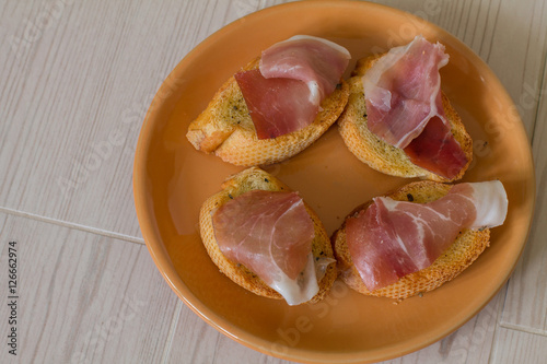 Four bruschettas with prosciutto and basil on orange plate on wood