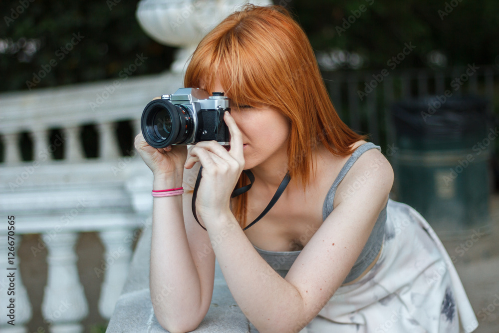 Young girl with orange hair taking pictures with an old camera retro style in a city park. She is happy to be able to use a reel camera again. 
