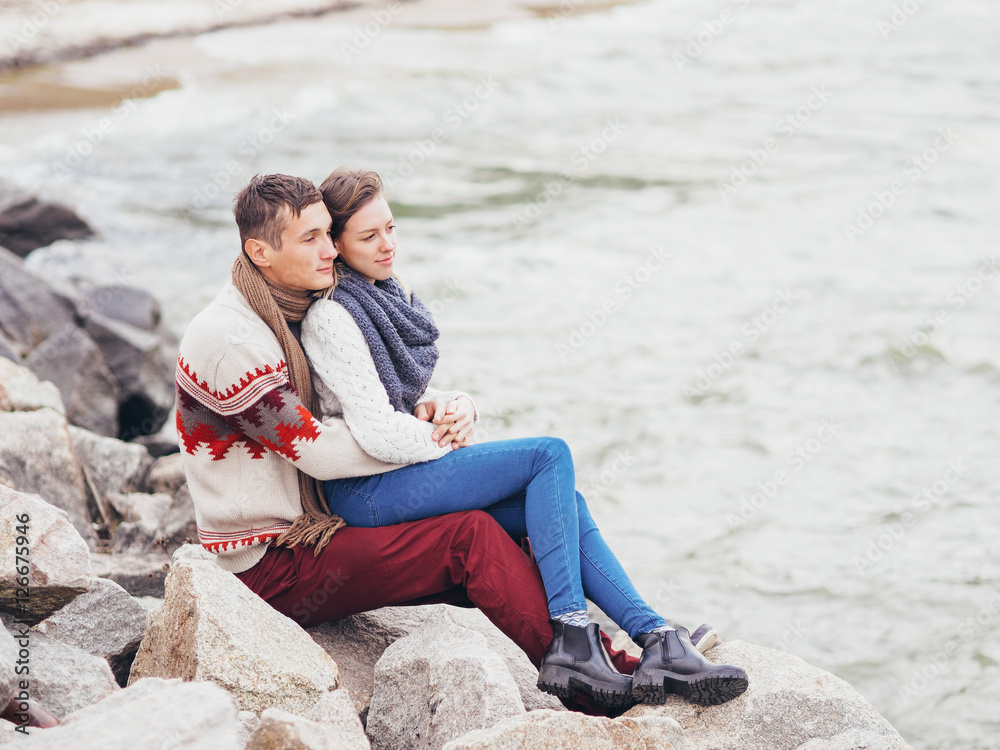 Young attractive couple in knitted sweaters on a cliff on sea shore, hugging and holding hands, creative toning