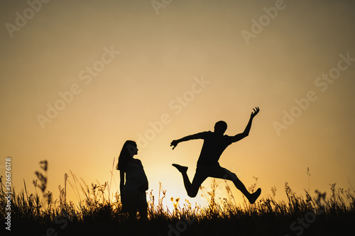 Silhouette portrait of funny young anonymous family in expectation of newborn. Future dad happily jumping over sunset sky background in evening in scenic nature landscape background. Happiness concept