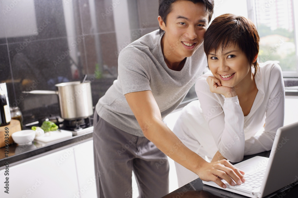 Couple in kitchen, using laptop, smiling at camera Couple in kitchen, using laptop, smiling at camera