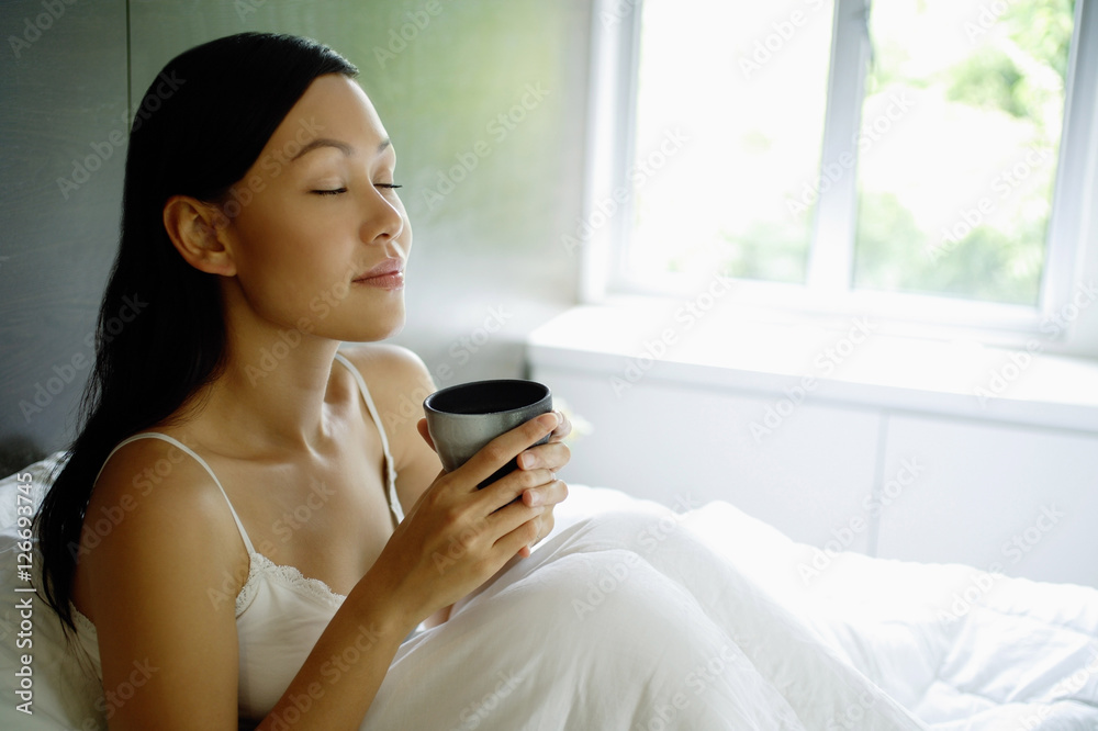 Woman sitting up in bed, holding cup, eyes closed Woman sitting up in bed, holding cup, eyes closed