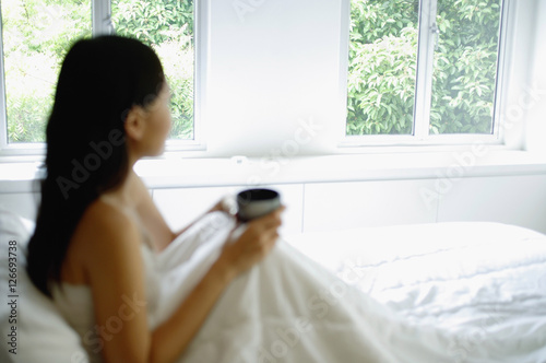 Woman sitting up in bed, holding cup, looking away