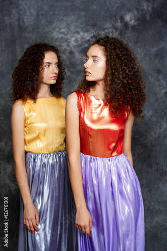 Beautiful sisters twins in bright dresses posing over grey background.
