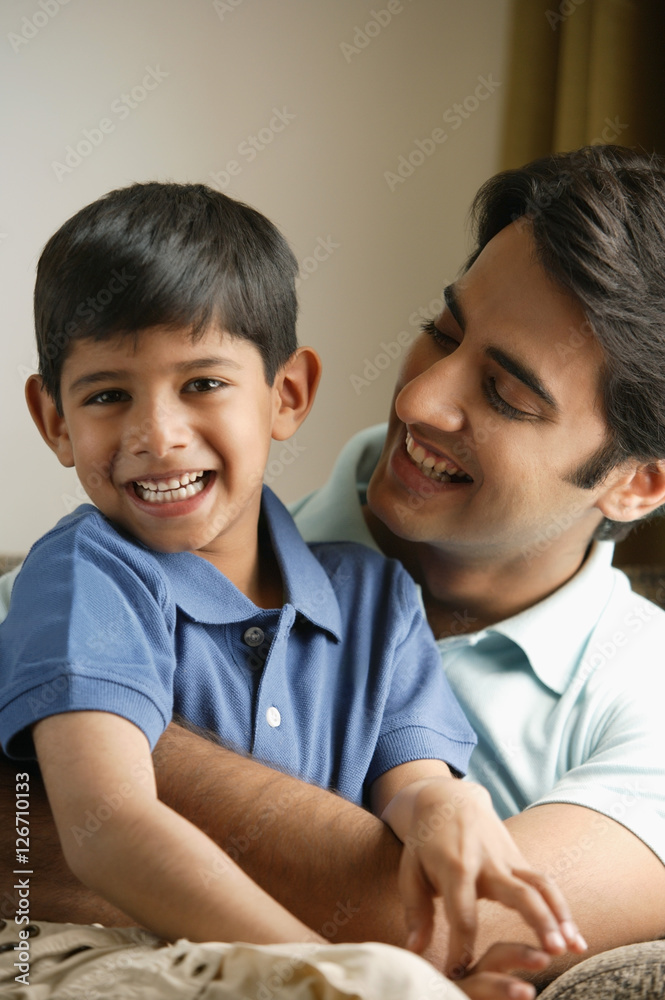 father hugging son on couch, son smiles at camera father hugging son on couch, son smiles at camera