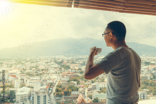 Asian man holding cup of coffee at office he looking out window. Cityscape backdrop. Selective focus and soft flare filter.