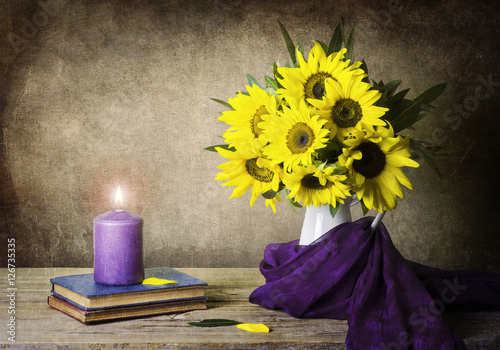 Fototapeta Naklejka Na Ścianę i Meble -  A classic still life with beautiful sunflowers placed in white jar which wrapped in purple scarf and candle on vintage books on rustic wooden table..