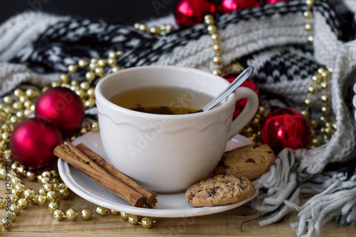 Horizontal photo of hot green tea in a white mug with spoon, cinnamon sticks, chocolate cookies, knitted blanket and christmas decoration