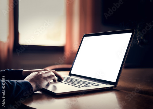 Man's hands using laptop with blank screen on desk in home dark interior.