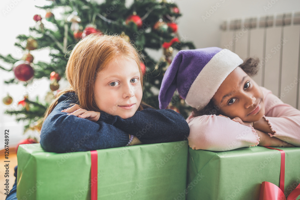 African and redhead little girls with gifts together in front of the Christmas tree. Natural light. 