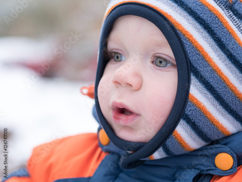 Attractive baby boy playing with the first snow. He smiles and looks snowman. Thick blue-orange jumpsuit bright striped hat on a year-old child.