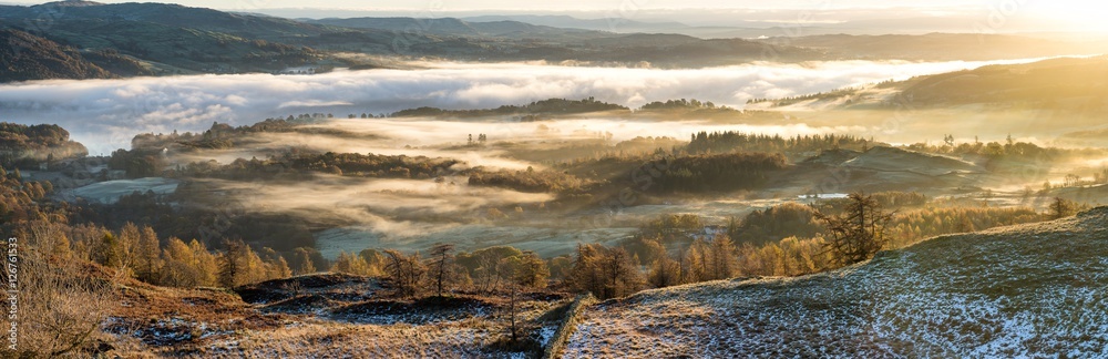 Fototapeta premium Vibrant golden sunlight shining over hills covered in Autumnal mist in the Lake District.
