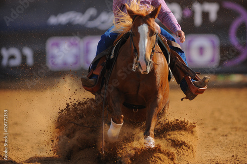 The front view of a rider in cowboy chaps and boots on a horseback running ahead and sliding the horse in the dirt