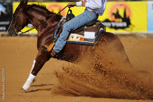 The side view of a rider in cowboy chaps and boots on a horseback running ahead and sliding the horse in the dirt