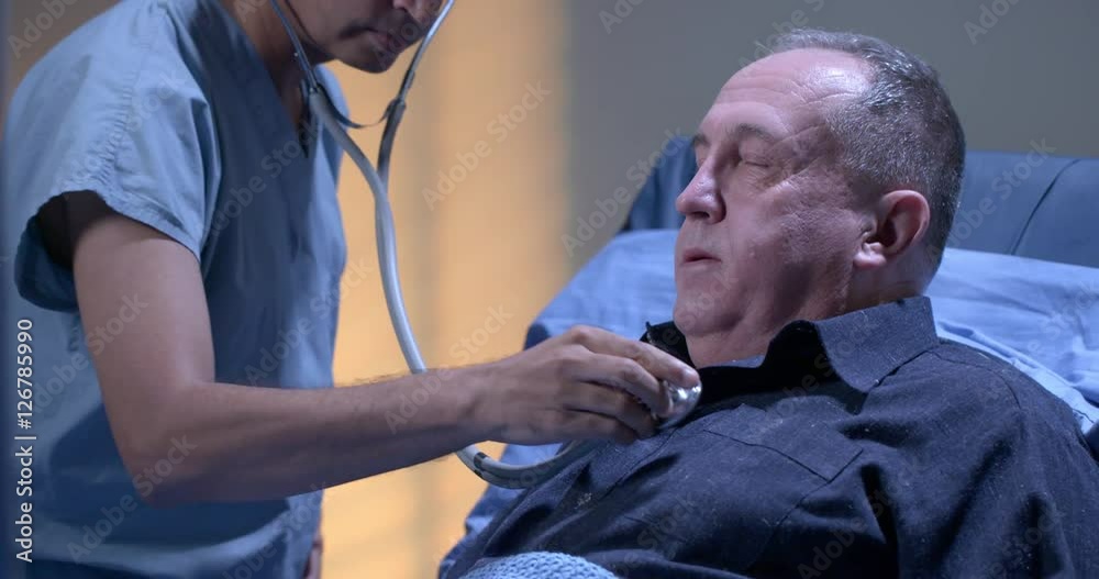 Male doctor uses stethoscope to check on mature male patient in hospital ward in the evening.  Medium close up recorded at 60fps