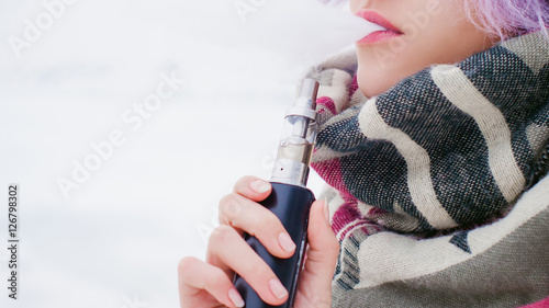 vaping girl. winter street portrait of a woman hipster, purple-dyed hair, a gray knitted hat and scarf. woman smokes an electronic cigarette in the street near the snow-covered river photo