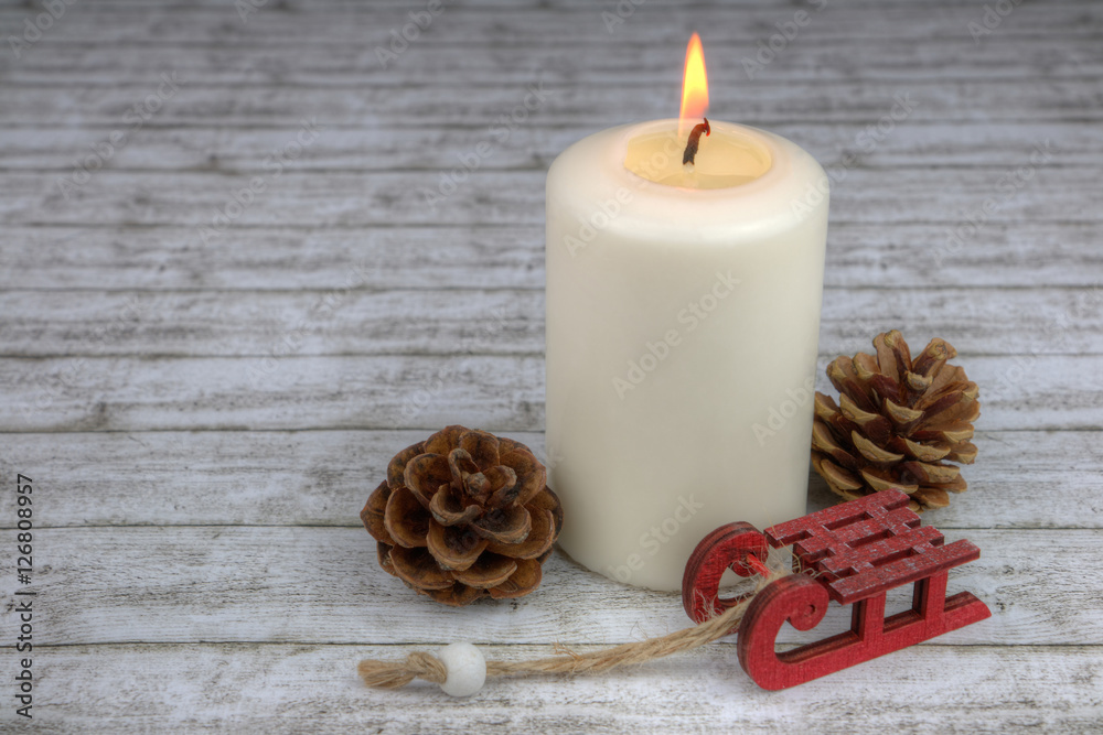 roter Schlitten und Tannenzapfen mit brennender weißer Kerze auf hellem Holz