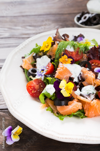 Fresh salad with smoked salmon, black olives, cherry tomatoes and edible flowers on wooden background.