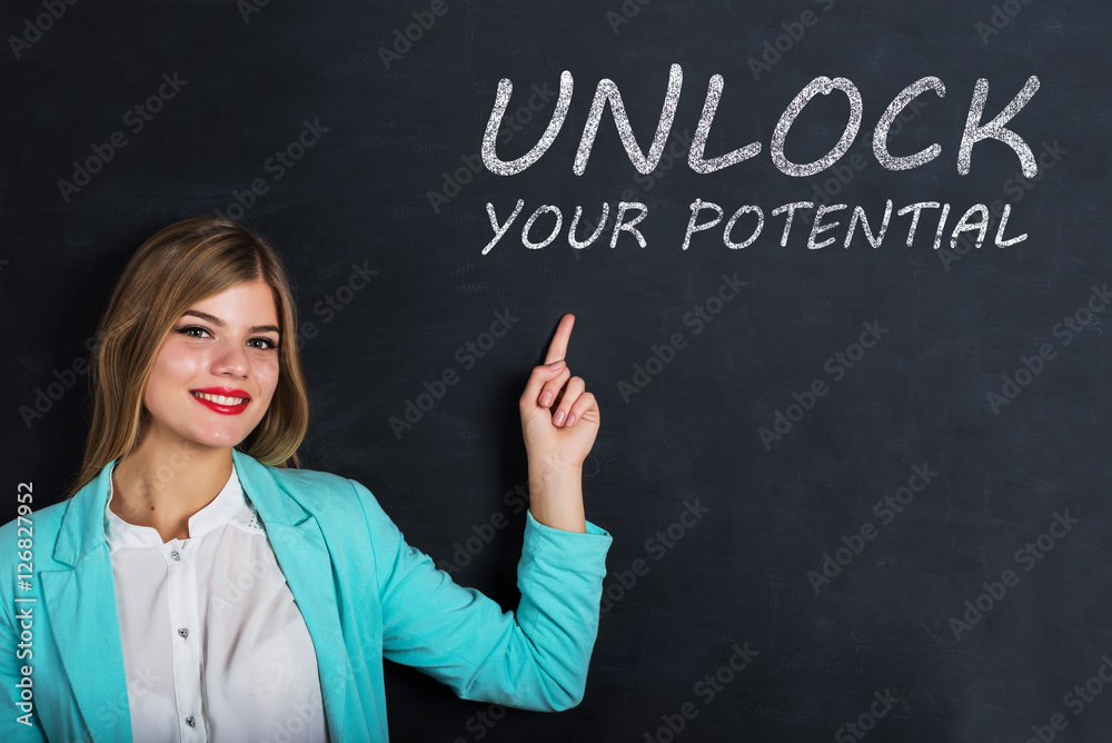 Beautiful young female student is pointing, looking at camera and smiling, standing against blackboard with text 