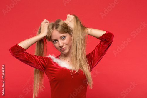 Beautiful young woman in Santa costume playing with a strand of her long silky hair, looking at camera on red background