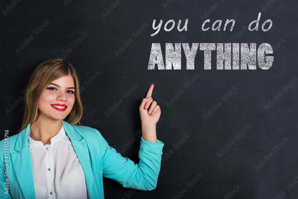 Beautiful young female student is pointing, looking at camera and smiling, standing against blackboard with text Beautiful young female student is pointing, looking at camera and smiling, standing against blackboard with text