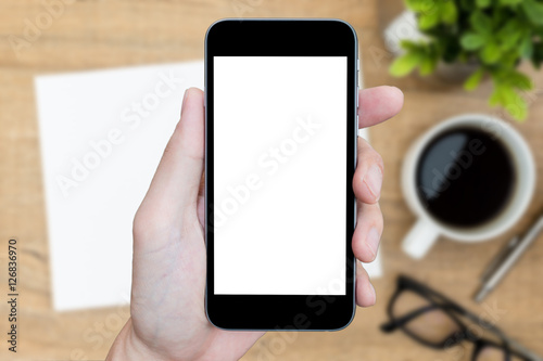 Hand is holding smartphone with blank mockup screen over the office desk.