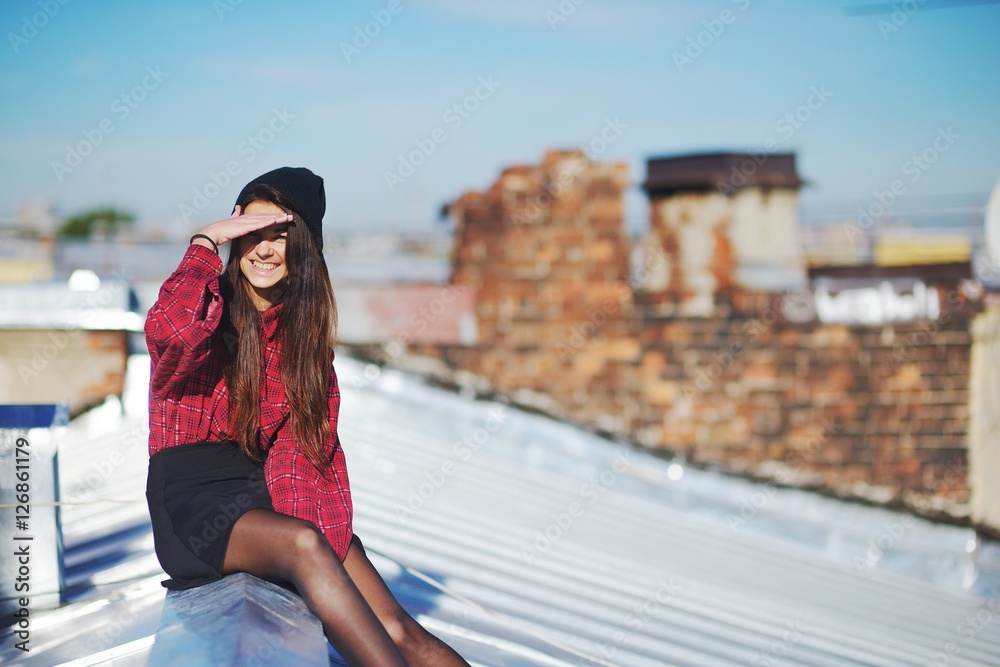 Happy smiling long-haired brunette girl in a red plaid shirt and short skirt sitting on the roof and hand protects the eyes from the bright sun against a brick wall.