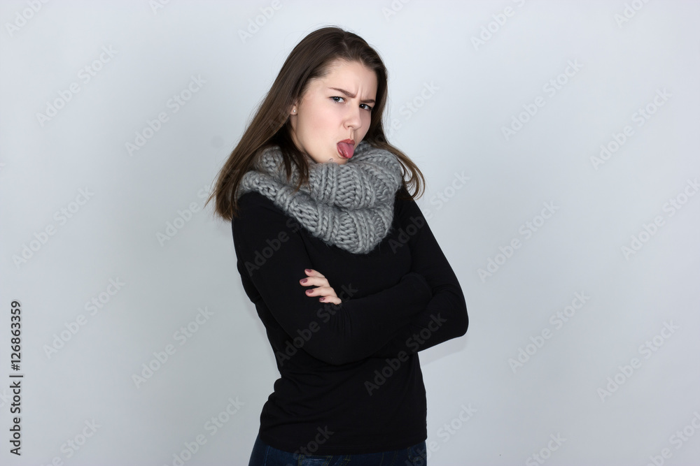 Portrait of a pretty offended sad gir in turtleneck sweater with scarf over gray background looking at camera and showing tongue Portrait of a pretty offended sad gir in turtleneck sweater with scarf over gray background looking at camera and showing tongue