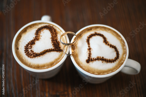wedding rings and two cups of coffee with foam in the form of heart. Wooden background.