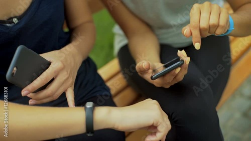 Two young ladies sitting on a bench checking fitness results on a smart phone. They wearing fitness trackers on arms.
