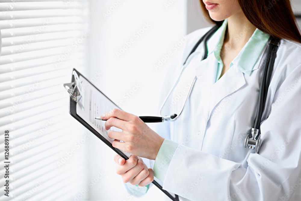 Female doctor filling up medical history standing near the window in hospital Female doctor filling up medical history standing near the window in hospital