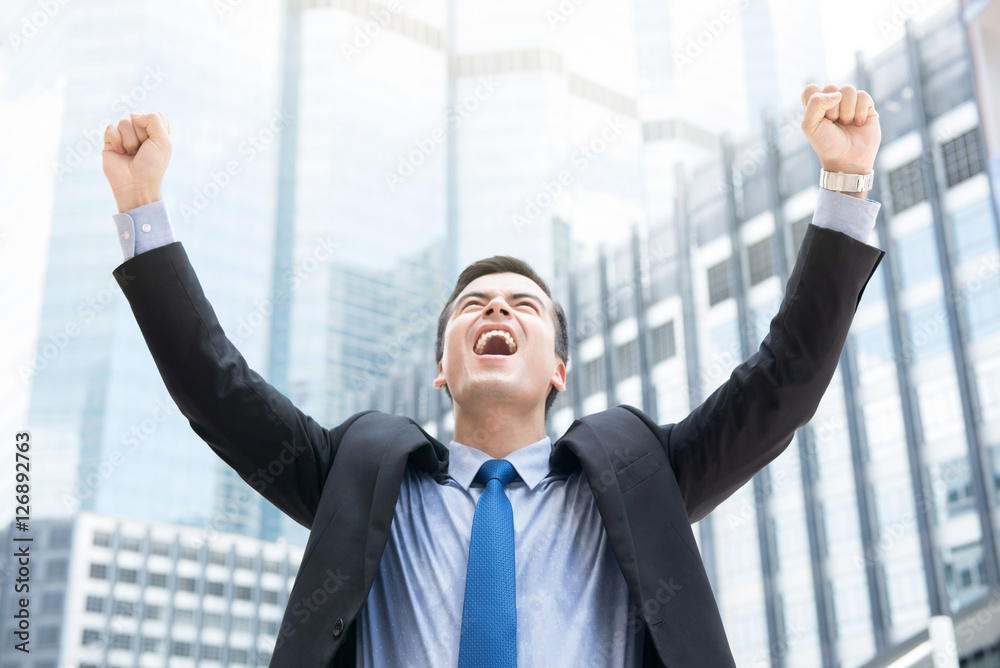 Businessman raising his fists in the air on blur office building background Businessman raising his fists in the air on blur office building background