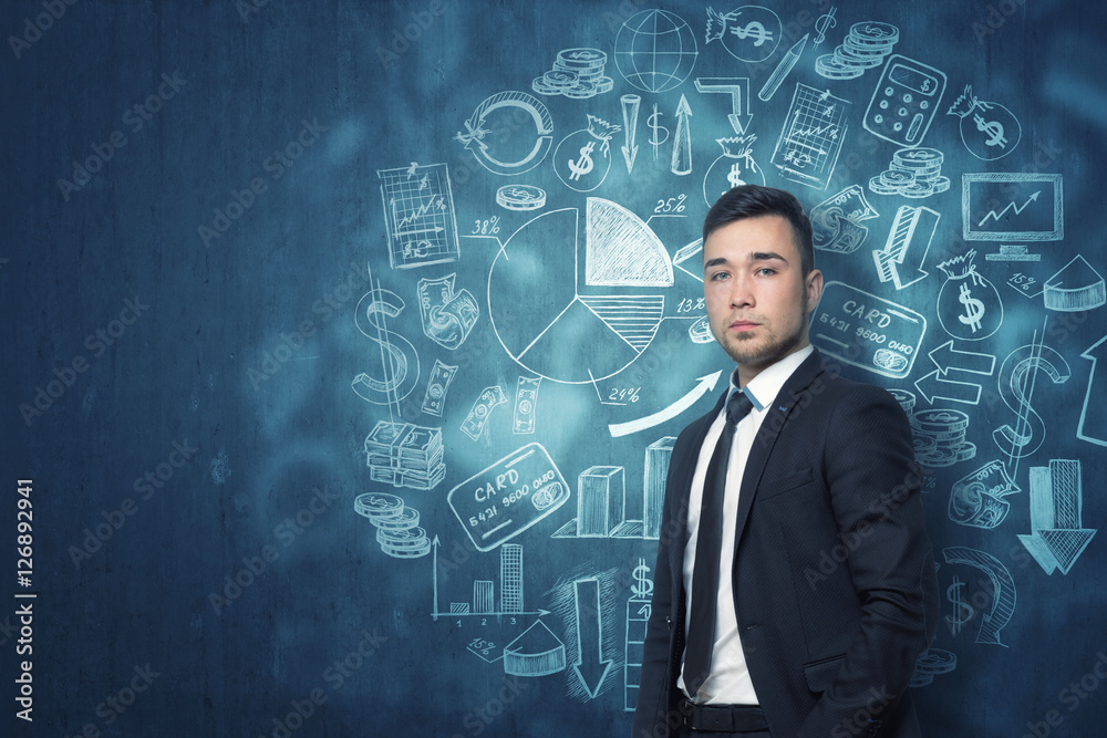 Young businessman standing in front of dark blue wall with different financial sketches Young businessman standing in front of dark blue wall with different financial sketches