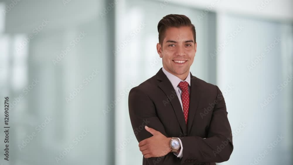 Happy smiling young Hispanic businessman standing in office with arms crossed looking at camera. Copyspace or copy space next to business professional with smile on his face Happy smiling young Hispanic businessman standing in office with arms crossed looking at camera. Copyspace or copy space next to business professional with smile on his face
